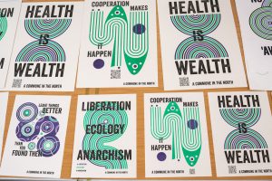 Photograph of various posters by A Commune in the North, saying things like 'health is wealth', and 'liberation,ecology,anarchism'