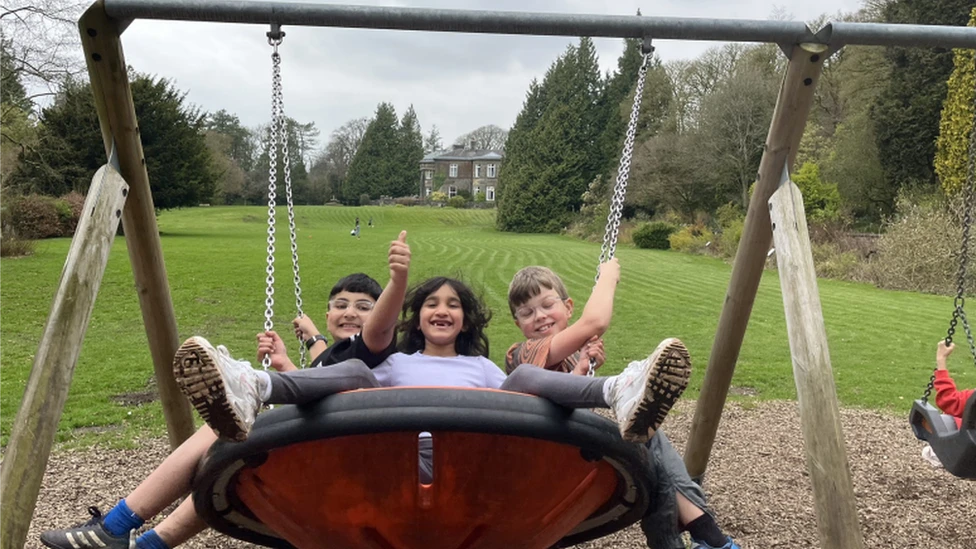 Three kids on a swing together, smiling and laughing. A lawn and a grand house in the background.